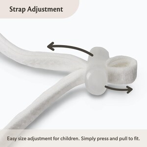Op de afbeelding: Een witte stoffen band met een doorzichtige plastic verstelknop. De afbeelding bevat de tekst Strap Adjustment en Easy size adjustment for children. Simply press and pull to fit, met donkere pijlen die de beweging van de verstelknop aangeven.