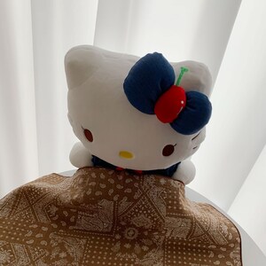 May include: A white plush character toy with a large dark blue bow featuring a red cherry accent. The character has black oval eyes and a yellow nose. It is partially covered by a brown fabric cloth with a white repeating paisley pattern.