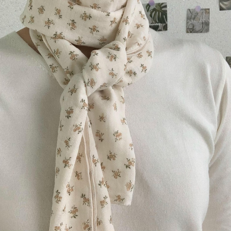 May include: A lightweight off-white scarf featuring a repeating pattern of small, muted brown floral clusters. The scarf is tied in a simple knot around the neck, resting against a plain, long-sleeved off-white top with a ribbed texture.