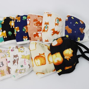 Op de afbeelding: Een collectie van tien stoffen mondkapjes met diverse Shiba Inu-hondenpatronen in kleuren zoals turquoise, zwart, marineblauw, perzik, cr&egrave;me en wit. Elk masker heeft witte of zwarte elastische oorlussen met verstelbare sluitingen voor een pasvorm.