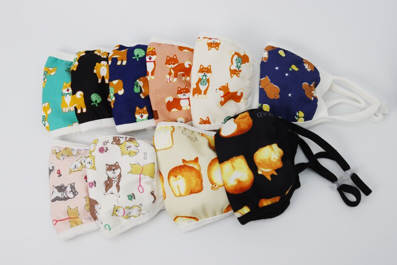 Op de afbeelding: Een collectie van tien stoffen mondkapjes met diverse Shiba Inu-hondenpatronen in kleuren zoals turquoise, zwart, marineblauw, perzik, cr&egrave;me en wit. Elk masker heeft witte of zwarte elastische oorlussen met verstelbare sluitingen voor een pasvorm.