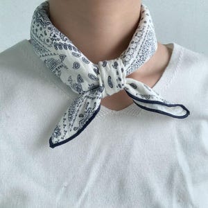 Puede incluir: Un pa&ntilde;uelo blanco con un estampado de cachemira azul oscuro atado al cuello. La tela tiene una apariencia suave y texturizada con un dobladillo azul oscuro en los bordes. El pa&ntilde;uelo se lleva sobre una camiseta de punto blanca de manga corta.