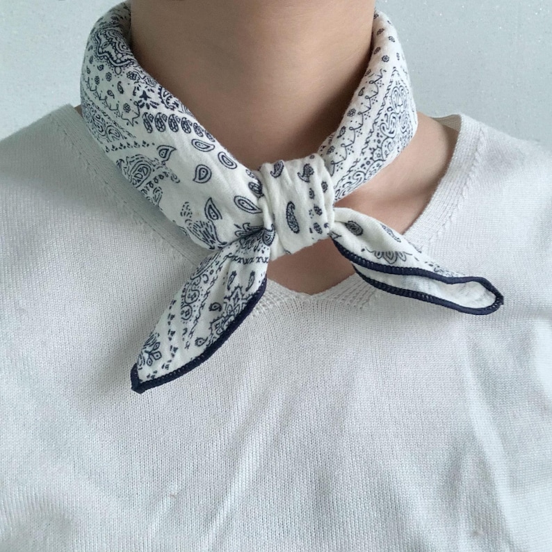 Puede incluir: Un pa&ntilde;uelo blanco con un estampado de cachemira azul oscuro atado al cuello. La tela tiene una apariencia suave y texturizada con un dobladillo azul oscuro en los bordes. El pa&ntilde;uelo se lleva sobre una camiseta de punto blanca de manga corta.
