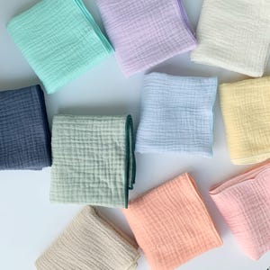 May include: A collection of ten square muslin cloths in various pastel and solid colours, including mint green, lavender, cream, navy blue, sage green, light blue, yellow, peach, beige, and soft pink, all featuring a textured, crinkled fabric weave.