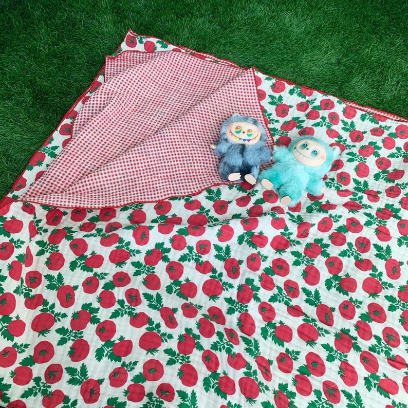 May include: A soft fabric blanket featuring a red tomato and green leaf pattern on a white background, with a red and white gingham underside. Two plush monster toys, one blue and one light green, are resting on the folded blanket.