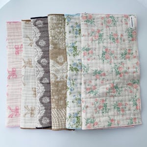 May include: A set of six rectangular cotton cloths featuring various patterns including pink bows, tan bows, a dark gray lace design, a tan floral print, blue flowers, and pink roses. Each cloth has a quilted texture and a small white label.