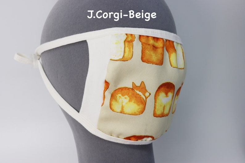 Op de afbeelding: Een beige mondkapje met een herhalend patroon van ge&iuml;llustreerde corgi-honden en broden. Het masker heeft witte stoffen randen en witte zijbanden. De tekst J.Corgi-Beige is in het wit boven het masker op de grijze achtergrond gedrukt.