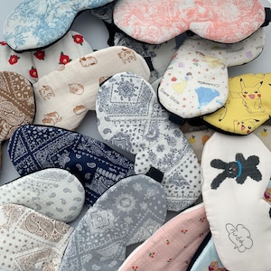 May include: Assortment of colorful sleep masks with various patterns and designs. Some masks feature cartoon characters like Pikachu and a bunny rabbit. Others have floral, paisley, and geometric patterns.