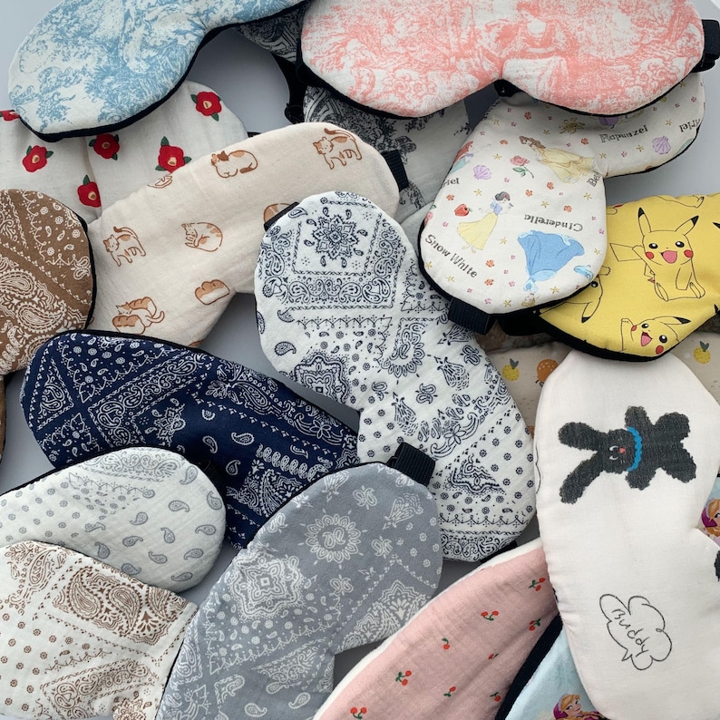 Op de afbeelding: Een assortiment kleurrijke slaapmaskers met verschillende patronen en ontwerpen. Sommige maskers hebben cartoonfiguren zoals Pikachu en een konijn. Andere hebben bloemen-, paisley- en geometrische patronen.
