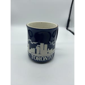 May include: A ceramic mug with a dark blue exterior and a cream-colored interior. The mug features a white silhouette of the Toronto skyline and the city's name. The handle is on the right side.