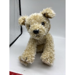 May include: A plush toy dog with light brown, curly fur. The stuffed animal has a black nose, dark eyes, and floppy ears. The dog is sitting upright, with its paws extended.
