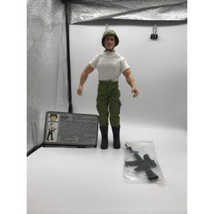 May include: A vintage action figure of a soldier, wearing a white t-shirt, green pants, and a green helmet. The figure has black boots and is standing next to a card that says "GRUNT". A plastic bag contains a black toy rifle.