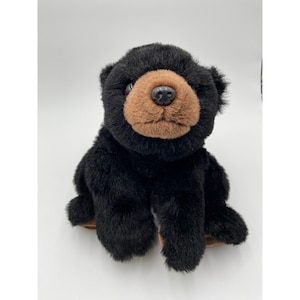 Può includere: Un orsacchiotto di peluche con morbida pelliccia nera che copre il corpo e la testa, con un muso color marrone chiaro a contrasto. L'orso ha un naso liscio in plastica scura e occhi rotondi e scuri incastonati nella pelliccia, in posizione seduta.