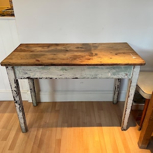 May include: A rectangular wooden table with a distressed finish. The tabletop is a natural wood colour, while the legs and apron are painted white with visible wear. The table measures approximately 122 cm long and 61 cm wide.