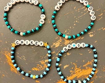 May include: Four beaded bracelets with white letter beads spelling out "MERCEDES", "YABADABADO", "CRIKEY", and "GR63". The bracelets feature black, turquoise, and light blue beads, with gold star accents on the "CRIKEY" bracelet.