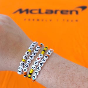 Peut inclure: Un poignet portant cinq bracelets en perles avec les mots "LETS GO LAND", "MCLAREN", "LALN 4", "PAPAYA" et "ON" écrits en lettres noires sur des perles blanches. Les bracelets sont faits de perles colorées orange, jaunes, noires et bleues.