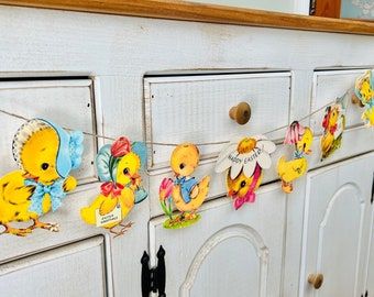 May include: A white painted cabinet with a string of Easter chick paper cutouts hanging from the top. The cutouts are yellow, blue, and pink with the text "Happy Easter!" on one of the chicks.