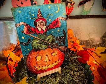 May include: Vintage Halloween postcard featuring a witch flying on a broomstick over a jack-o'-lantern. The witch is wearing a green dress and a pointed hat, and she has a black cat riding on her shoulder. The background is a blue sky with stars.