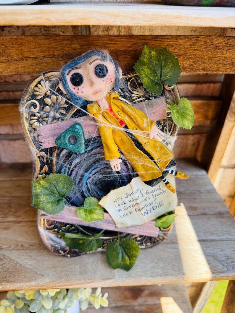 Peut inclure: Une poup&eacute;e Coraline aux cheveux bleus et aux yeux en boutons portant un imperm&eacute;able jaune, fix&eacute;e sur une plaque d&eacute;corative avec des feuilles vertes et un effet toile d'araign&eacute;e. Une note indique : Hey Jonesy. Look what I found in Gramma's trunk. Look familiar? WYBIE.