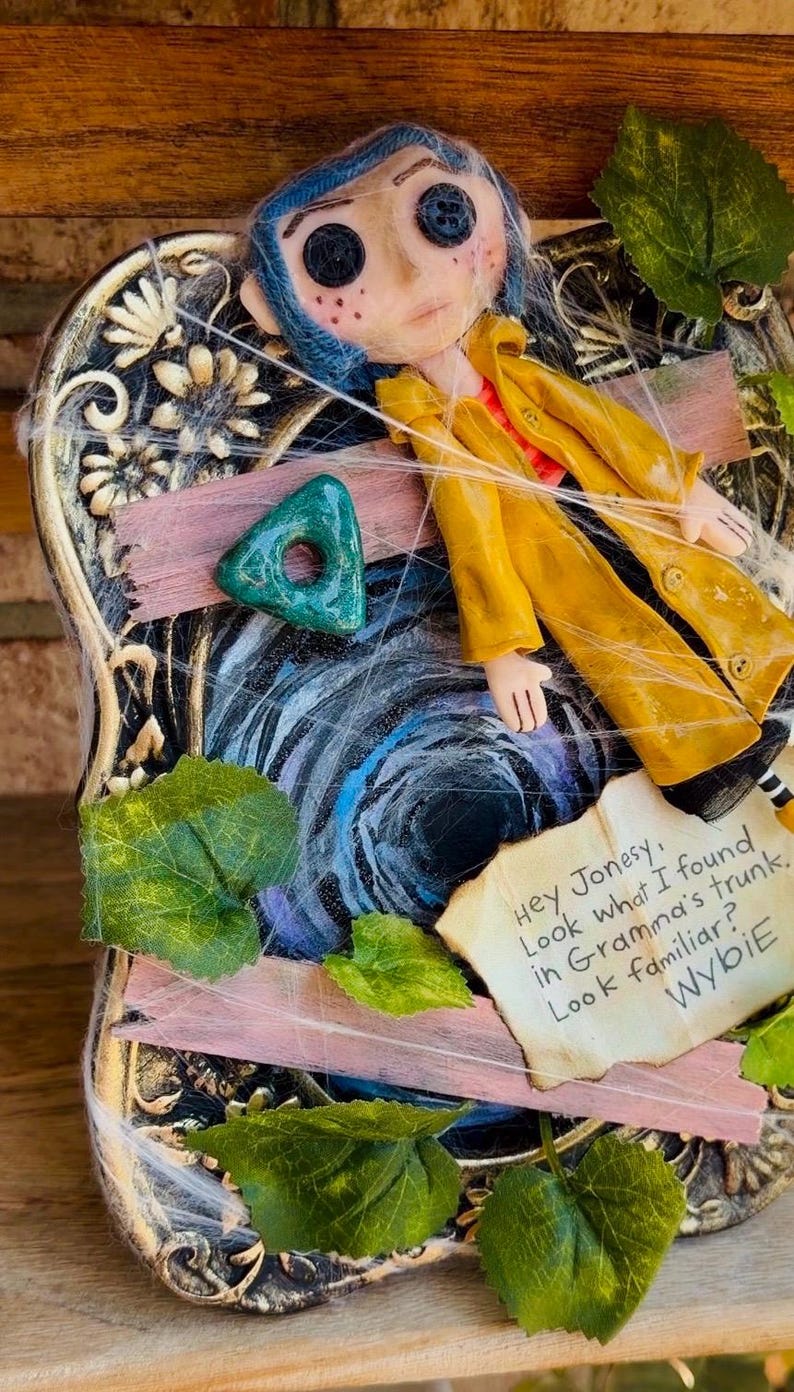 Peut inclure: Une poup&eacute;e aux cheveux bleus et aux yeux en boutons portant un imperm&eacute;able jaune, plac&eacute;e sur un cadre d&eacute;coratif avec une pierre triangulaire verte, des feuilles artificielles et une note : Hey Jonesy, Look what I found in Gramma's trunk. Look familiar? Wybie.