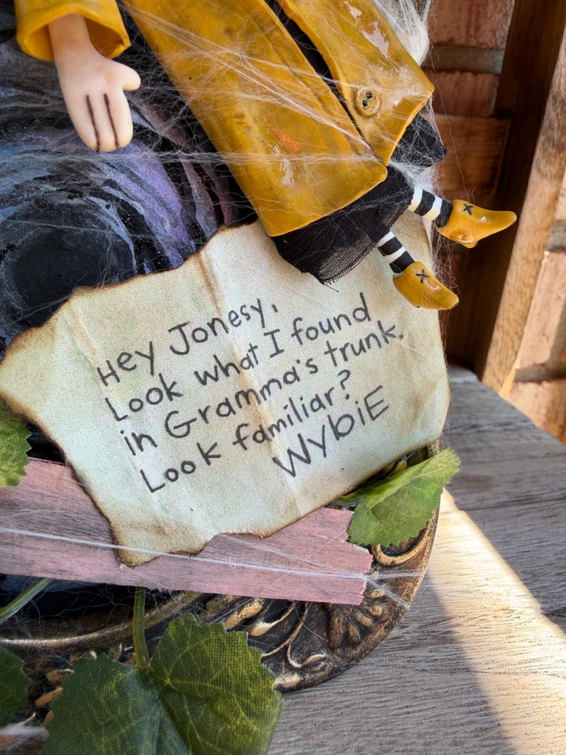 Peut inclure: Une figurine portant un imperm&eacute;able jaune et des chaussettes ray&eacute;es noires et blanches, tenant un papier avec le texte : Hey Jonesy, Look what I found in Gramma's trunk. Look familiar? Wybie. La figurine est d&eacute;cor&eacute;e de toiles d'araign&eacute;e artificielles.