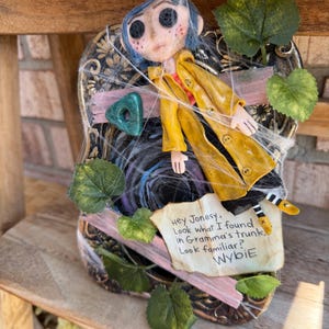 Peut inclure: Une poup&eacute;e Coraline aux cheveux bleus et aux yeux en boutons portant un imperm&eacute;able jaune, fix&eacute;e sur un cadre noir d&eacute;cor&eacute; de feuilles vertes, d'un ruban rose et de fausses toiles d'araign&eacute;e. Une note indique : Hey Jonesy, Look what I found in Gramma's trunk. Look familiar? Wybie.
