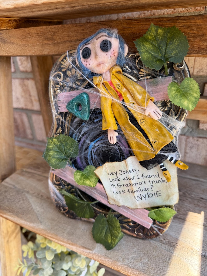 Peut inclure: Une poup&eacute;e Coraline aux cheveux bleus et aux yeux en boutons portant un imperm&eacute;able jaune, fix&eacute;e sur un cadre noir d&eacute;cor&eacute; de feuilles vertes, d'un ruban rose et de fausses toiles d'araign&eacute;e. Une note indique : Hey Jonesy, Look what I found in Gramma's trunk. Look familiar? Wybie.