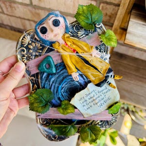 Peut inclure: Une plaque murale d&eacute;corative repr&eacute;sentant une figurine de Coraline aux cheveux bleus et aux yeux en boutons, v&ecirc;tue d'un imperm&eacute;able jaune. Elle inclut une pierre triangulaire verte, des feuilles artificielles, des toiles d'araign&eacute;e et une note : Hey Jonesy, Look what I found in Gramma's trunk. Look familiar? WYBIE.
