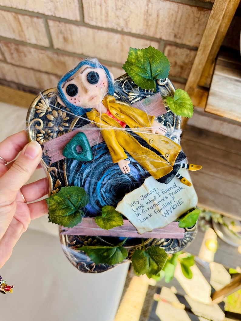 Peut inclure: Une plaque murale d&eacute;corative repr&eacute;sentant une figurine de Coraline aux cheveux bleus et aux yeux en boutons, v&ecirc;tue d'un imperm&eacute;able jaune. Elle inclut une pierre triangulaire verte, des feuilles artificielles, des toiles d'araign&eacute;e et une note : Hey Jonesy, Look what I found in Gramma's trunk. Look familiar? WYBIE.