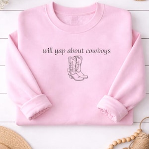 Könnte beinhalten: Hellrosa Sweatshirt mit dem Text "will yap about cowboys" über einer Strichzeichnung von Cowboy-Stiefeln. Das langärmlige Shirt hat einen Rundhalsausschnitt und gerollte Manschetten. Der Hintergrund ist eine weiße Holzoberfläche.