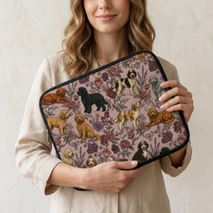 May include: A rectangular laptop sleeve featuring a pattern of various dog breeds and floral elements in shades of brown, black, and cream against a mauve background. The sleeve has a black zipper and is held by a person.