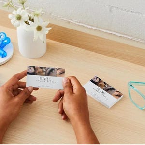 May include: A business card with the text "BARE Beauty Studio" and eye images is held on a wooden surface. A small white vase with flowers, blue clips, and teal glasses are also visible.