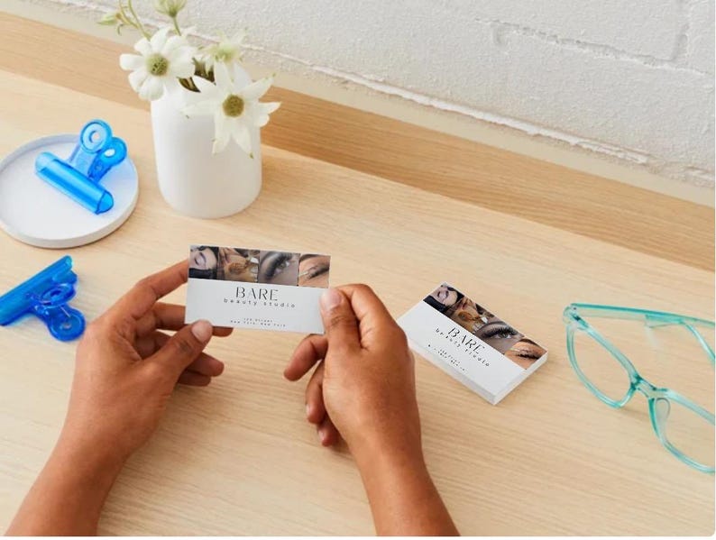 May include: A business card with the text "BARE Beauty Studio" and eye images is held on a wooden surface. A small white vase with flowers, blue clips, and teal glasses are also visible.