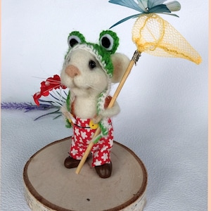 May include: A white felt mouse wearing a green frog hat and red and white overalls holds a butterfly net with a yellow net and a blue butterfly. The mouse is standing on a wooden base.