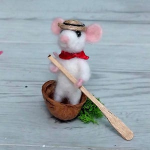 May include: A white felt mouse wearing a straw hat and a red scarf is sitting in a walnut shell and rowing with a wooden paddle.
