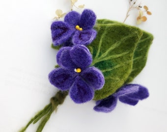 May include: A felt flower bouquet with three purple flowers and a green leaf. The flowers have yellow centers.