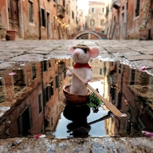 Peut inclure: Une figurine de souris en feutrine blanche portant un petit chapeau de paille et une écharpe rouge, assise dans un bateau en coquille de noix. La souris tient une longue rame en bois fine, naviguant sur une flaque d'eau sur des pavés.