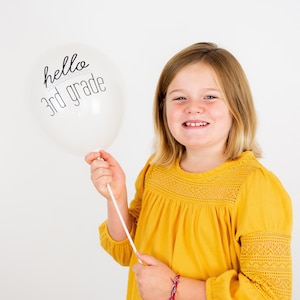 Könnte beinhalten: Ein junges Mädchen hält einen weißen Ballon mit dem Text "hello 3rd grade" in Schwarz.