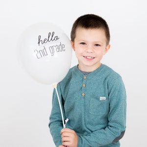 May include: A white balloon with the text "hello 2nd grade" in black lettering. A young boy is holding the balloon and smiling at the camera.
