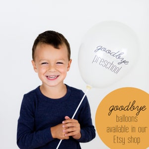 May include: A young boy smiles while holding a white balloon with the text "goodbye preschool" printed on it. A yellow circle in the background says "goodbye balloons available in our Etsy shop".