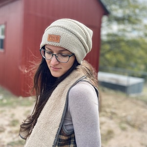 Puede incluir: Un gorro de punto beige con un parche de cuero marrón que dice "Pecu Leather Co."