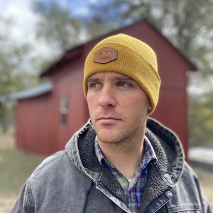 Könnte beinhalten: Ein Mann trägt eine senfgelbe Mütze mit einem braunen Lederpatch, auf dem "Explore" steht. Er trägt eine graue Jacke über einem karierten Hemd.