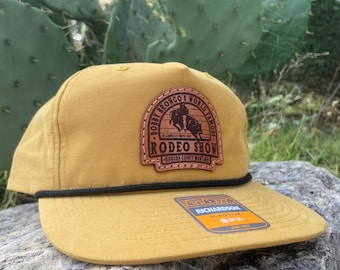 Puede incluir: Gorra de béisbol color mostaza con visera plana y detalle de cuerda negra. La gorra presenta un parche de cuero con el texto "Bobby Broncho's World Famous Rodeo Show" y un gráfico de un jinete a caballo. La etiqueta de la marca Richardson es visible.
