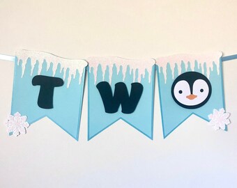 Pode incluir: Faixa azul e branca com a palavra "DOIS" e uma ilustração de pinguim. A faixa tem um floco de neve em cada extremidade e pingentes de gelo na parte superior.