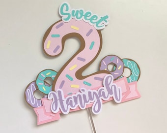 May include: A colorful birthday cake topper featuring a large number 2 designed as a donut with pink frosting and sprinkles. The topper includes the word "Sweet" and a banner with the name "Haniyah". Additional donut decorations complete the festive design.