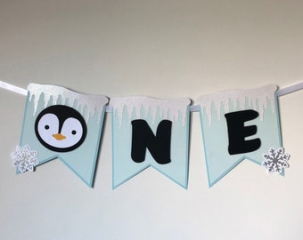 Pode incluir: Uma faixa azul-clara com três bandeirolas presas a uma fita branca. As bandeirolas exibem um rosto de pinguim e as letras O, N e E em preto. Cada bandeirola é decorada com pingentes de gelo com glitter prateado e flocos de neve.