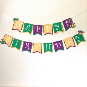 May include: A colorful banner with the words "Happy 1st Birthday" in white letters on a purple, green, and gold background. The banner is decorated with Mardi Gras masks and jester hats.