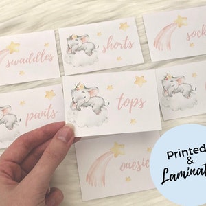 May include: Set of 8 baby shower gift tags featuring a pink and yellow watercolor design with a sleeping elephant and a shooting star. The tags are labeled with the following items: swaddles, pants, tops, onesie, shorts, socks, bibs.