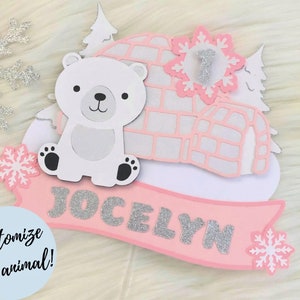 May include: Pink and white cake topper with a polar bear, igloo, and snowflakes. The name "Jocelyn" is written in silver glitter on a pink banner.