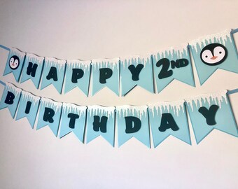 Pode incluir: Uma faixa de duas fileiras com bandeirolas azul-claras em formato de cauda de andorinha e detalhes de pingentes de gelo brancos com glitter no topo. As letras pretas formam HAPPY 2ND BIRTHDAY, com dois rostos de pinguim nas extremidades.
